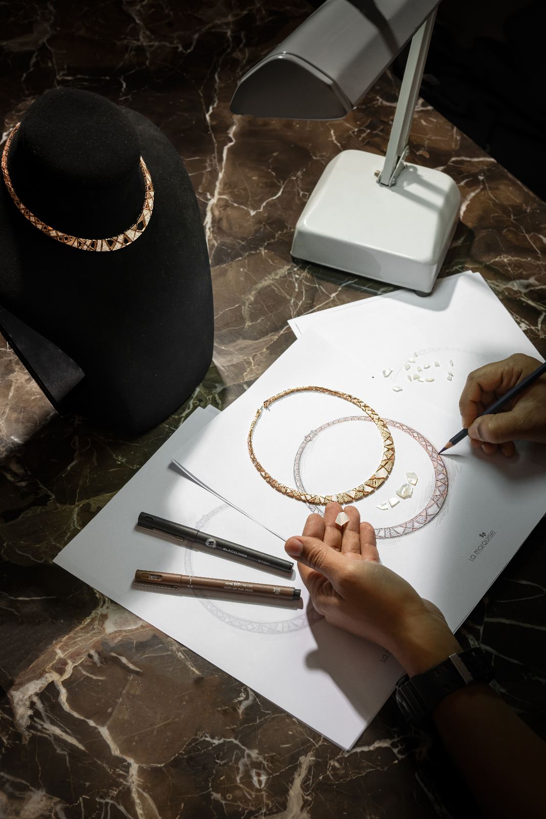 Jeweller sketching a custom necklace design with loose diamonds under an atelier lamp